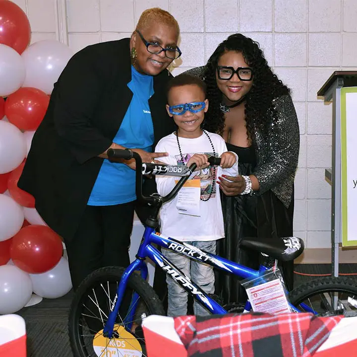 Anna’s Cupboard Founder & CEO and a Board Member, stand next to a young boy excited to receive a new bike