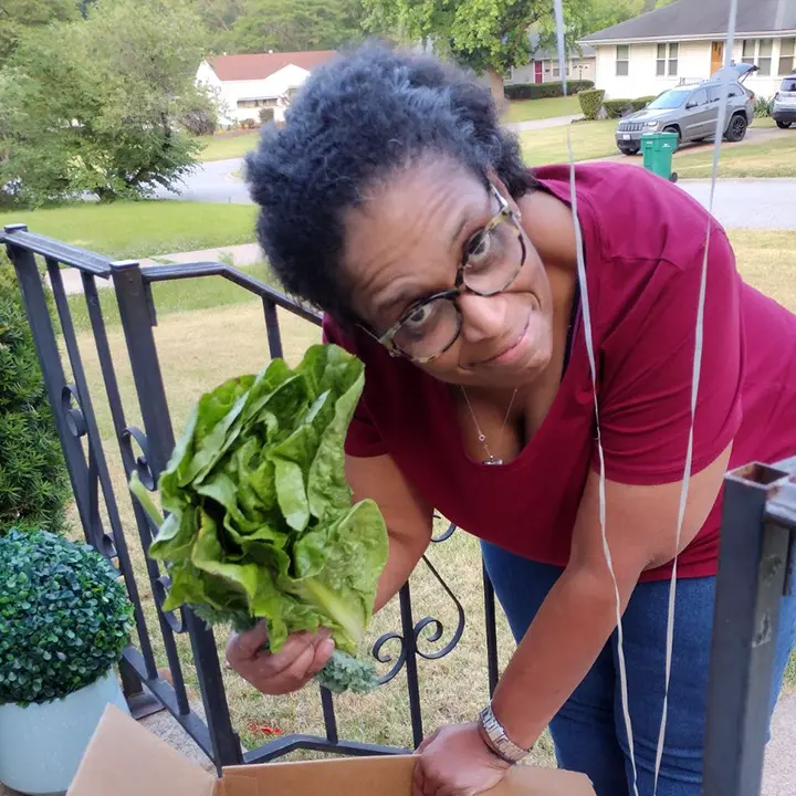 On a sunny, Spring day, a client of Anna’s Cupboard holds a head of lettuce after opening a box of freshly-dropped-off groceries